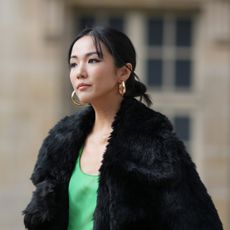 Yoyo Cao wears large golden earrings, a black faux fur fluffy long winter coat, a low-neck green dress, dark green tights, a green leather Givenchy bag, high heels shoes, outside Givenchy, during the Womenswear Spring/Summer 2024 as part of Paris Fashion Week on September 28, 2023 in Paris, France