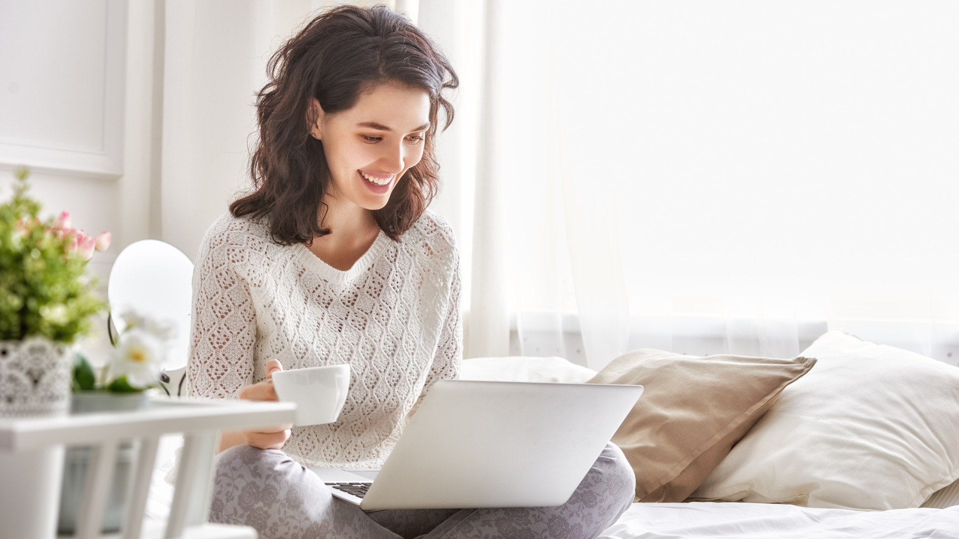 Wanita bahagia duduk di tempat tidur dengan kopi dan laptop