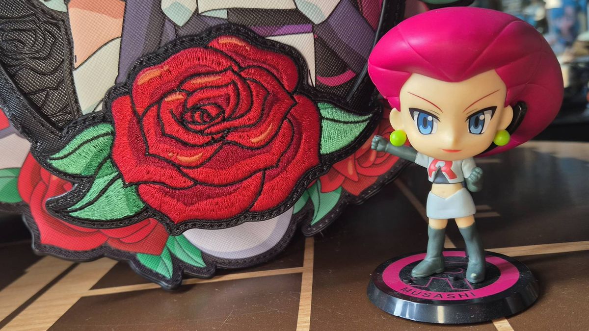 Close up of the rose on the front of the Team Rocket Cameo Mini Backpack with a Jessie toy to its side.