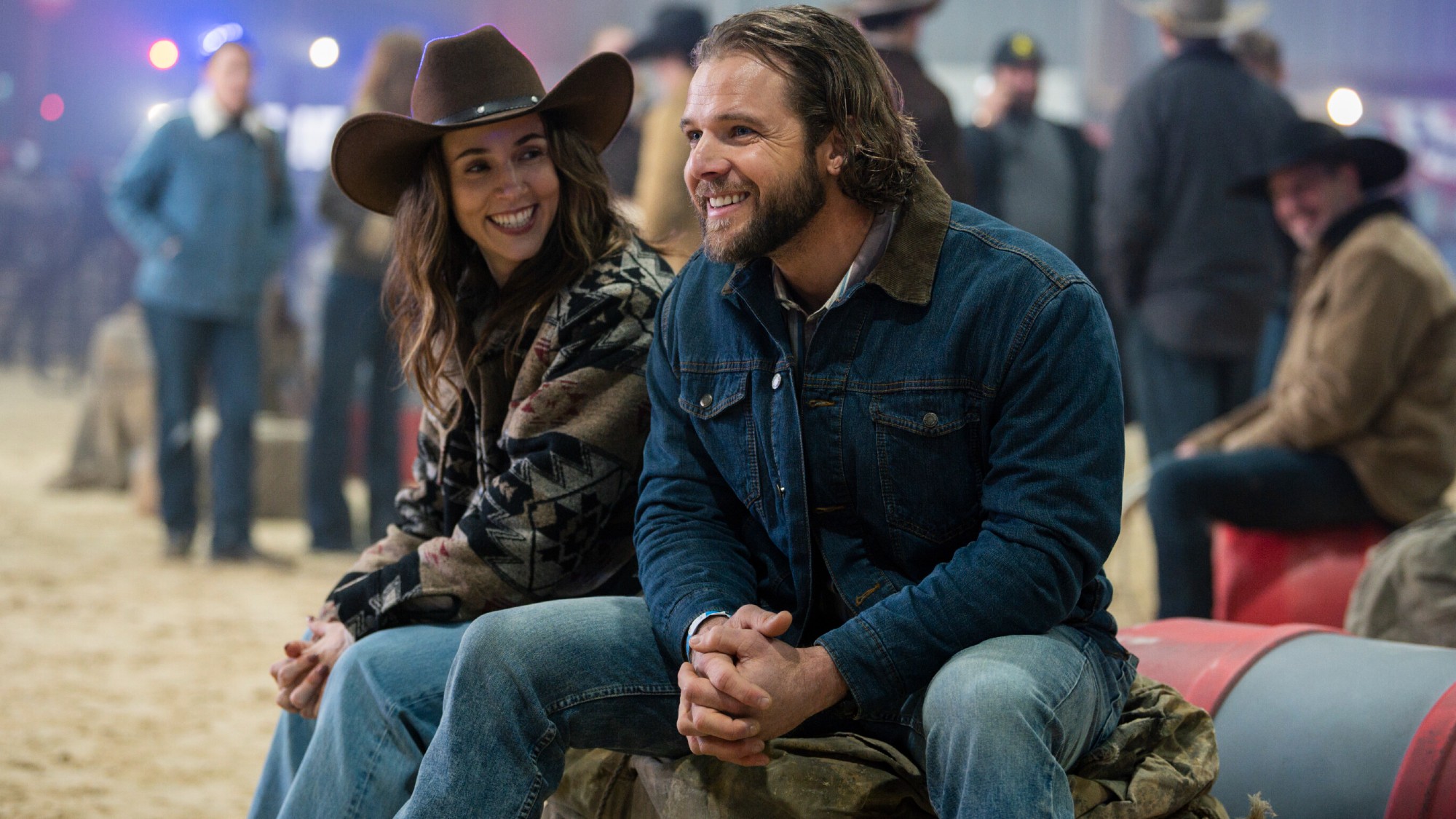 From left to right: Alona Tal and Max Thieriot sitting at a rodeo, they're both leaning against their knees and clasping their hands together. 