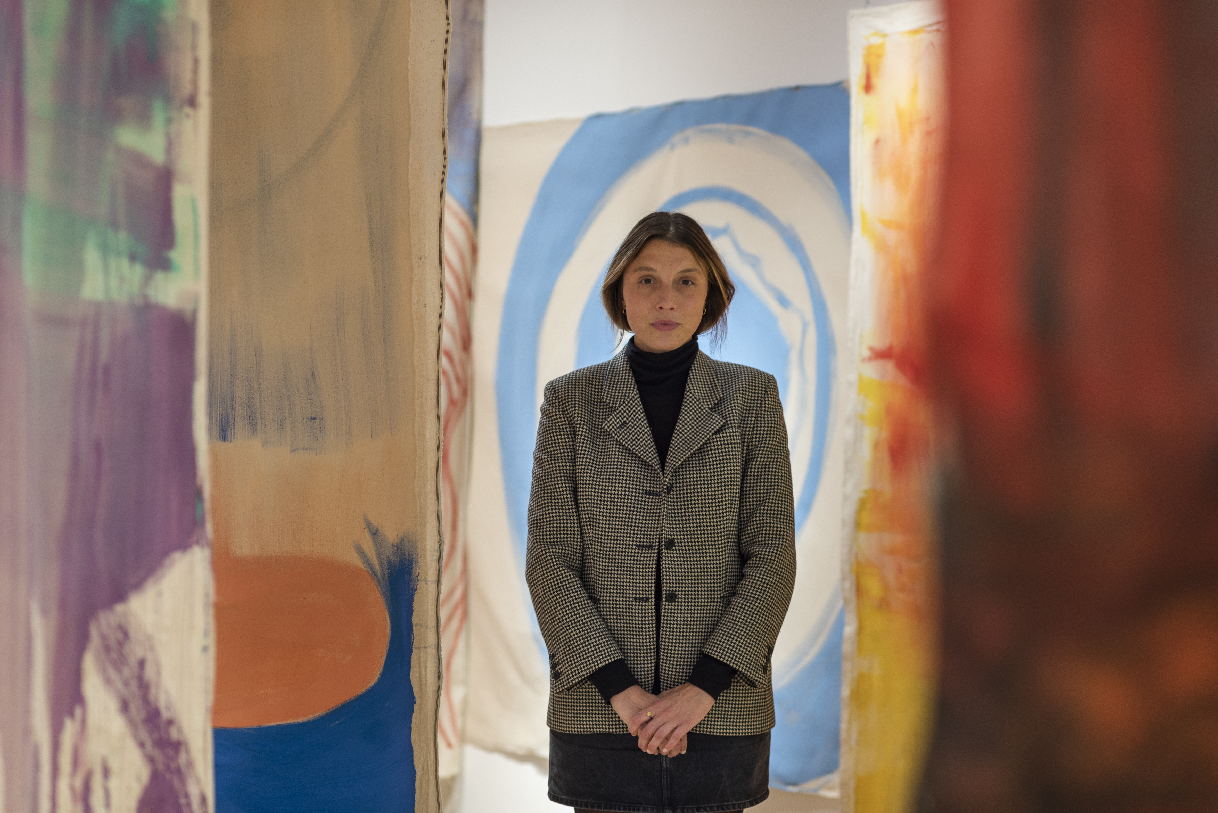 A young woman stands surrounded by handpainted, floating textiles in abstract motifs while wearing a dark turtleneck and a checkered white and black blazer.