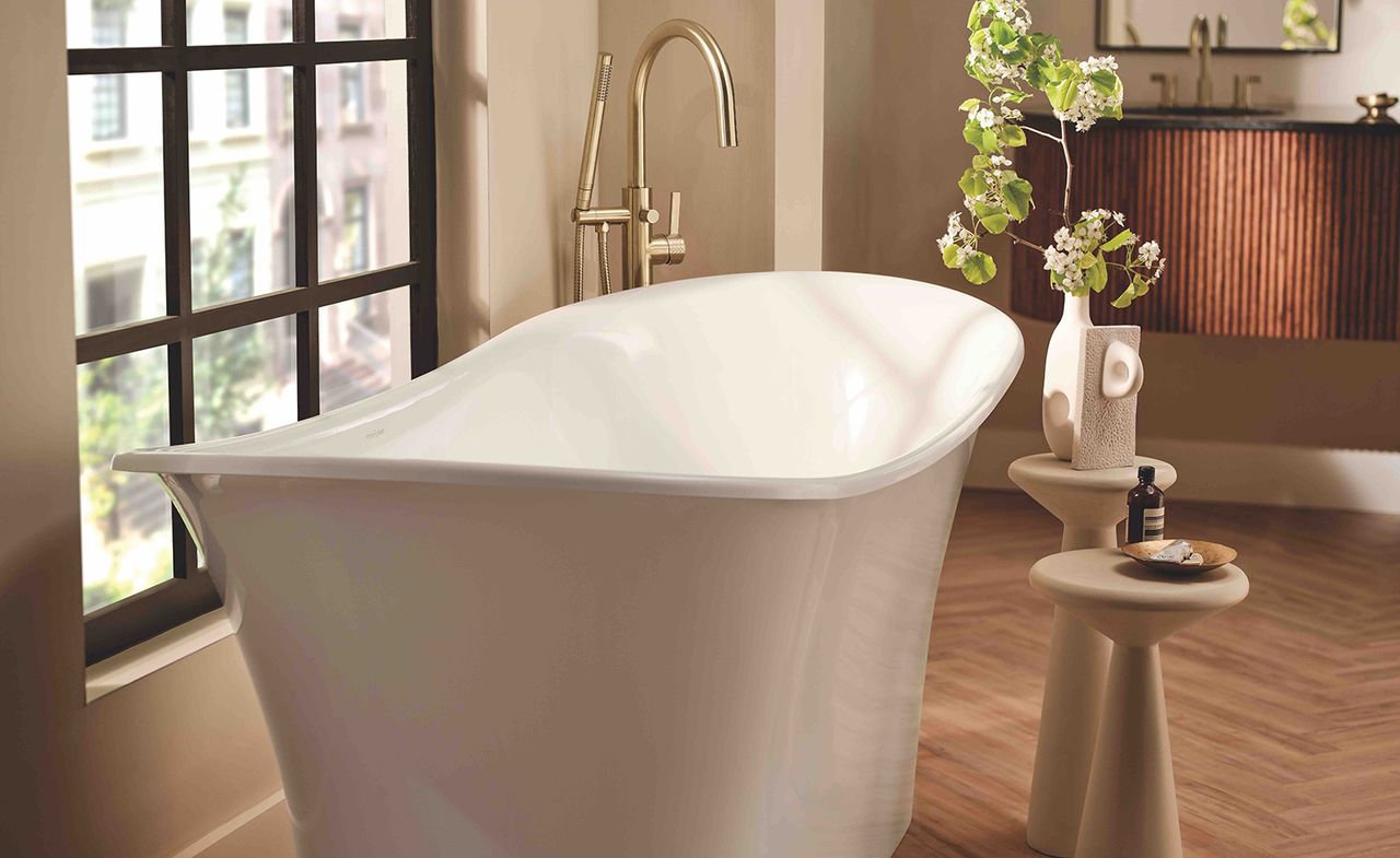 Bathroom image, wooden floor, white free stadning bathtub, gold taps, small side tables, black framed window with blurred view of surrounding area, white vase with flowers, neutral walls, wall mirror, brown ribbed design unit with gold taps