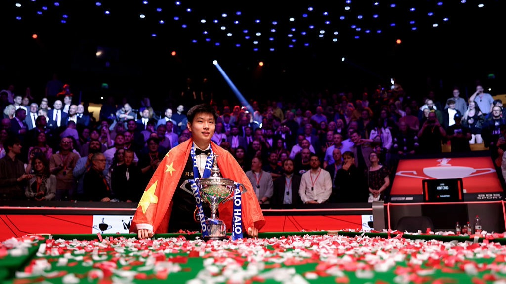 Zhao Xintong, wrapped in the flag of China, poses with the World Snooker Championship trophy after winning the 2025 tournament.