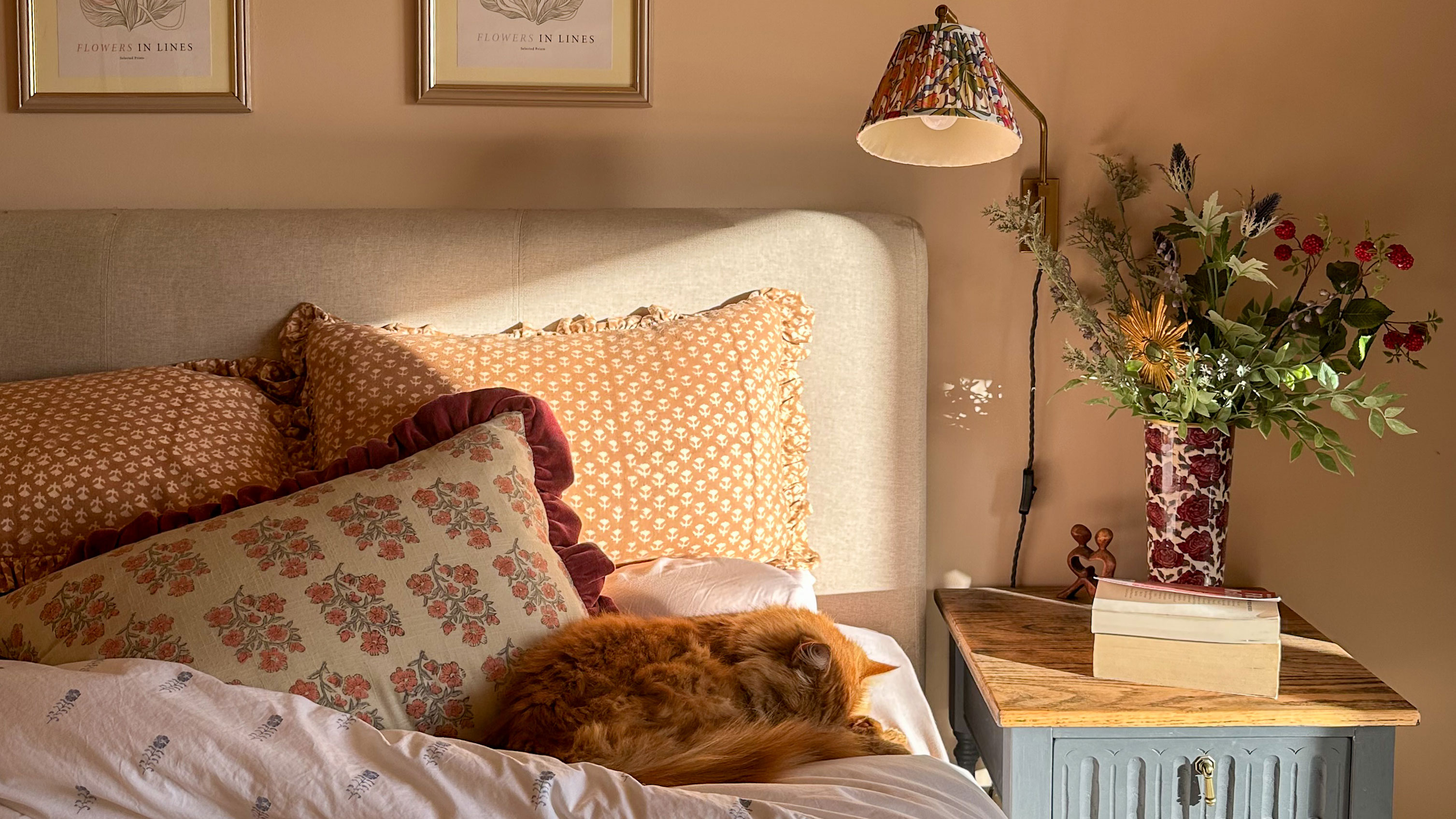 Bed with patterned cushions, cat curled up on duvet and vase of flowers on bedside table