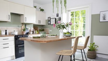 Kitchen with cream units, wood work surface, patterned floor tiles, green painted walls, rattan bar stools and plant in hanging planter