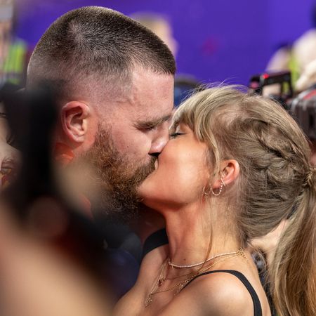 Taylor Swift has a ponytail and red lipstick and kisses NFL football player Travis Kelce as cameras film them