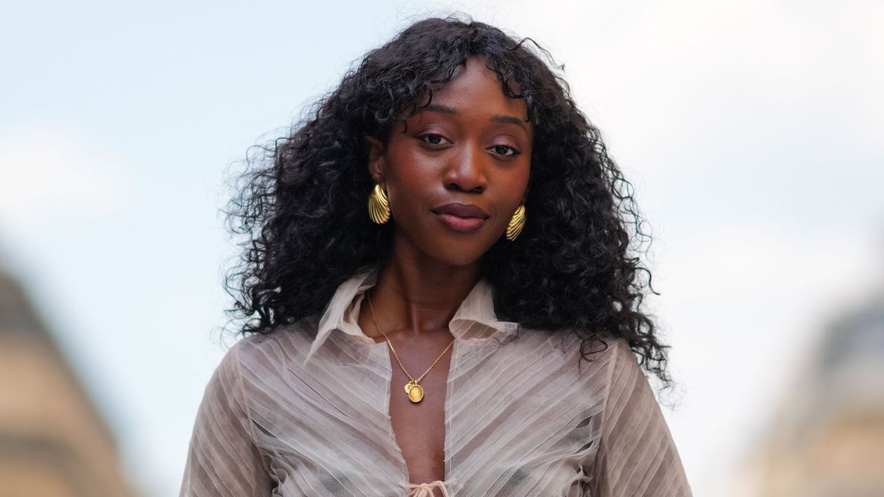 black woman with plum make-up and glowing nourished skin and gold earrings and sheer shirt