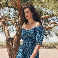 A woman walking past a tree wearing an off-the-shoulder blue floral Apricot dress with matching earrings