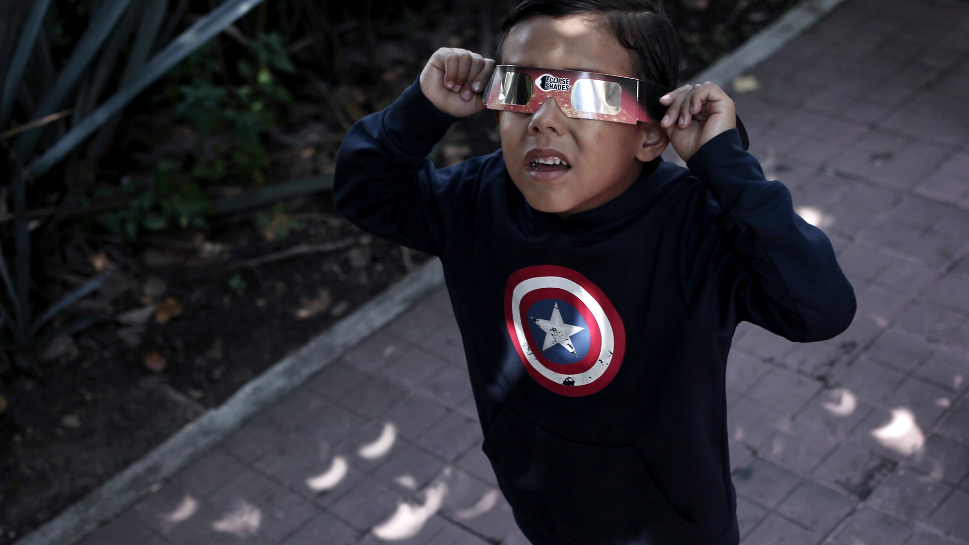 A child wearing eclipse glasses looks upward, with crescent shadows on the pavement below