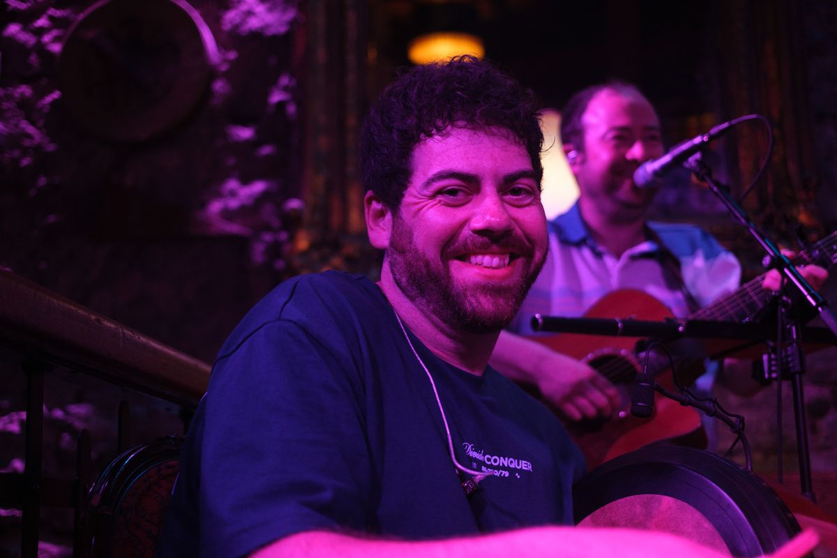 A smiling percussionist sits under purple stage lighting while a guitarist performs behind him.