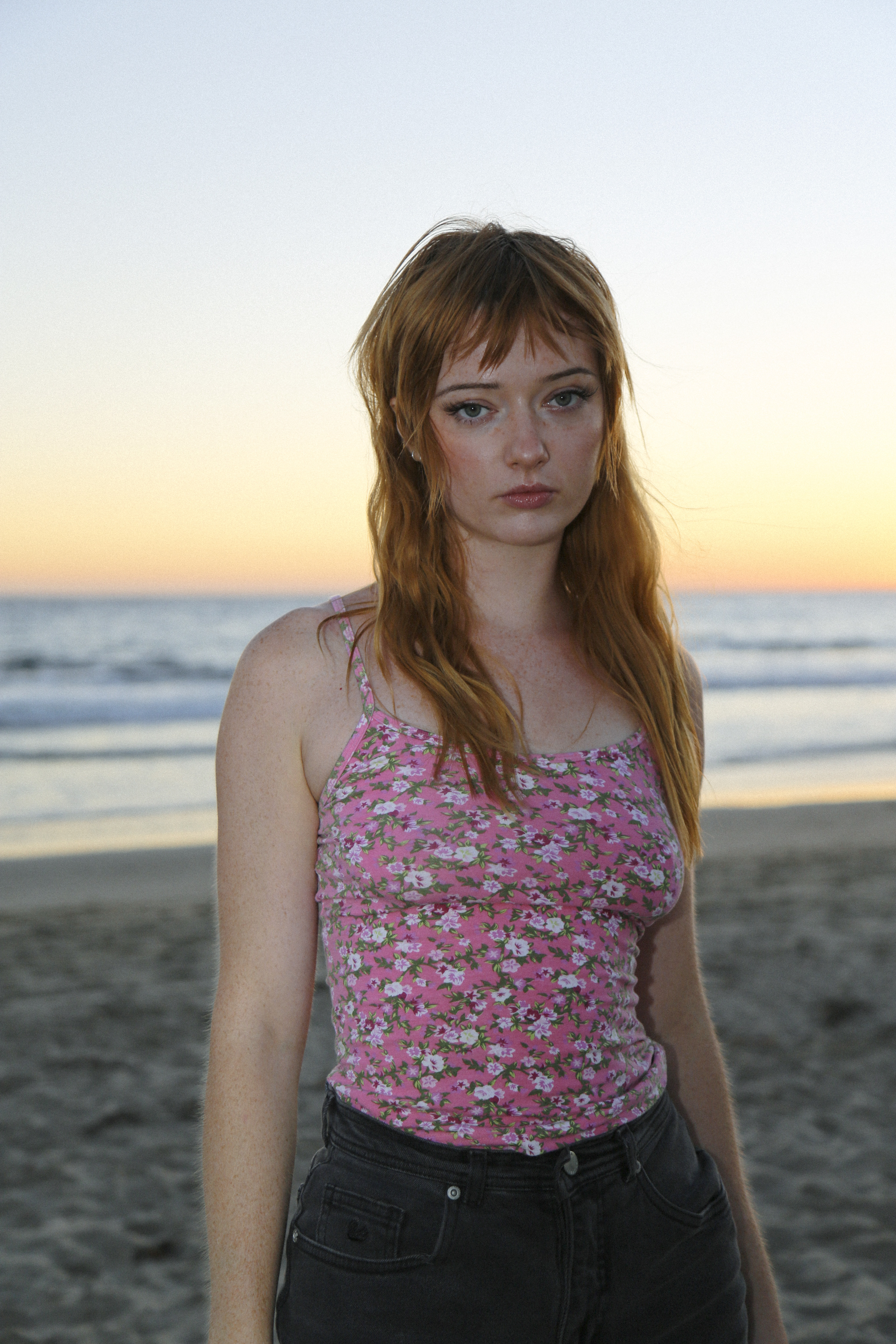 actress lindsey normington poses on a beach at sunset wearing a printed floral pink top and black denim shorts