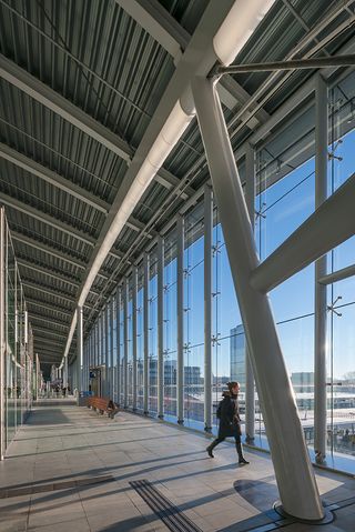 A walk way in the station featuring floor to ceiling glass open windows with views across the train tracks and city