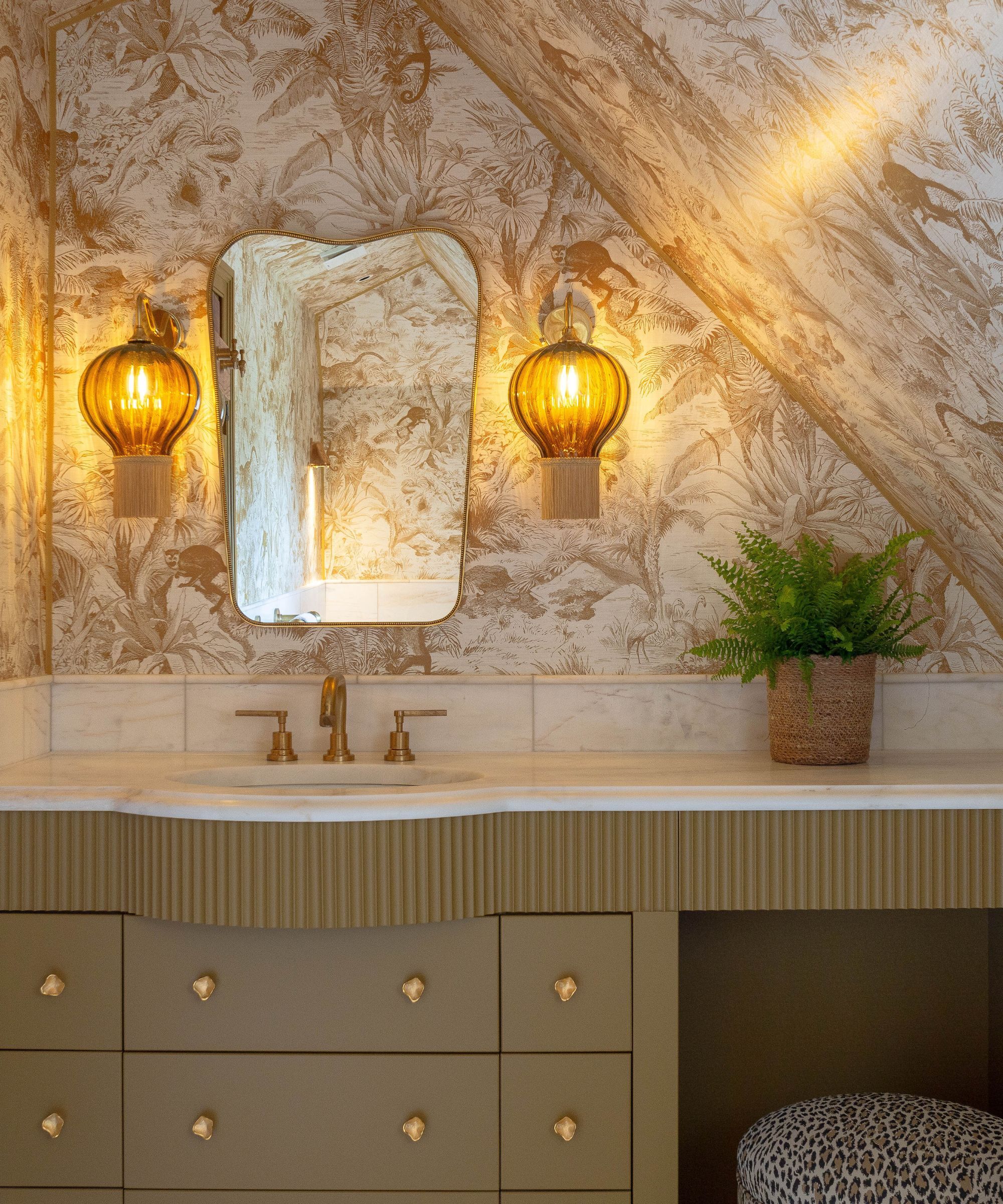 A bathroom vanity with a potted plant on top. An art deco mirror on the wall with two amber wall scone lights either side