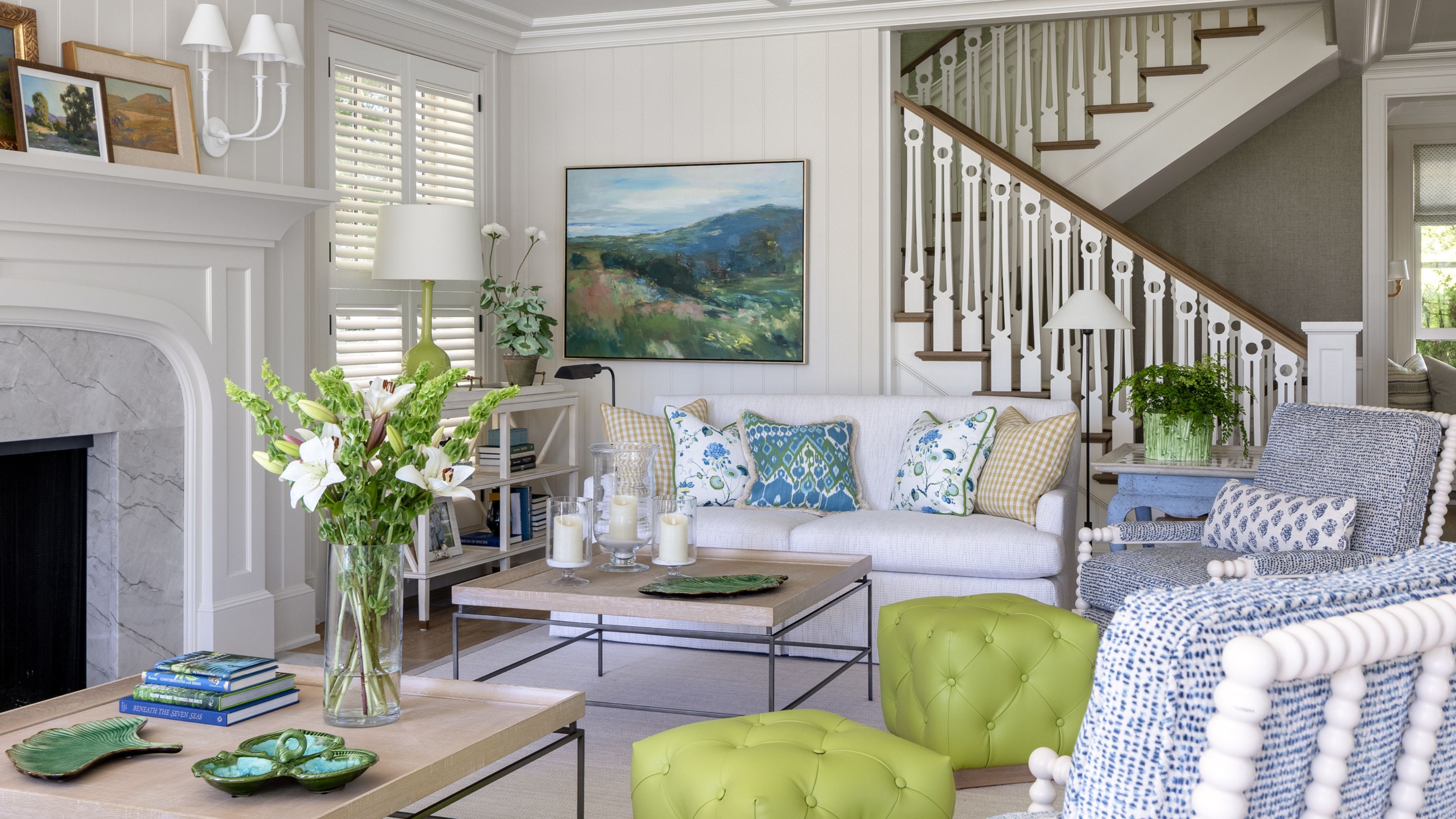 a light and bright blue and green styled summer living room with white couch, marble fireplace, green stools and blue accent chairs with white bobbin details