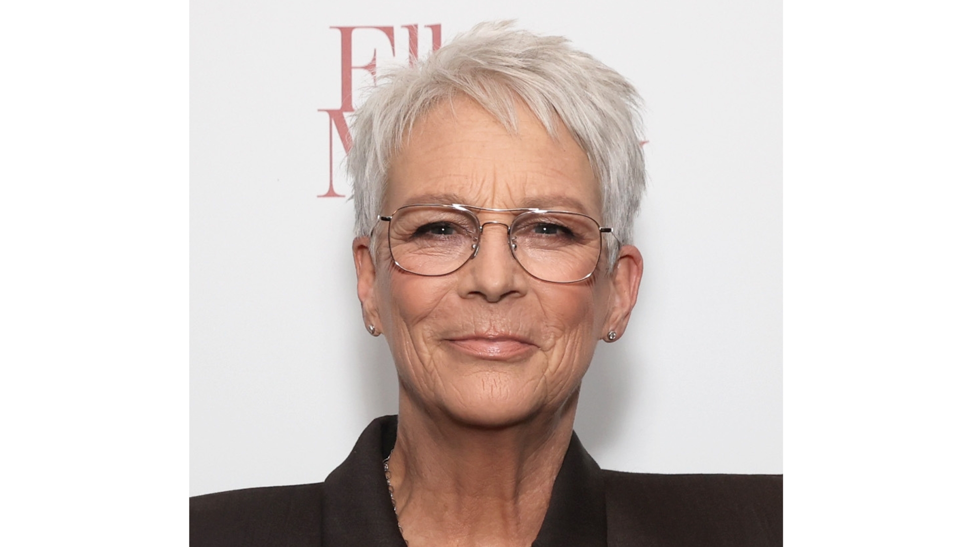 Image of Jamie Lee Curtis smiling with a grey pixie cut, wearing a black blazer and glasses