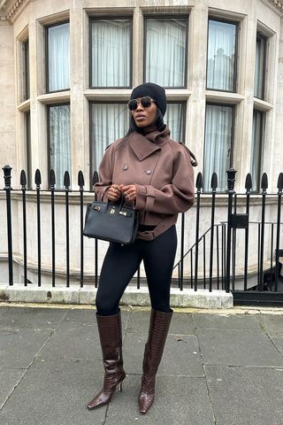 A woman wearing a black beanie, sunglasses, a brown peacoat, black leggings, brown knee-high boots, and a black handbag.