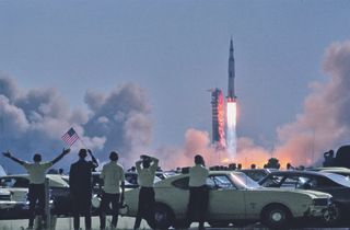 Spectators in a parking lot near the Vehicle Assembly Building wave an American flag as Apollo 11 begins its journey to the moon. The Saturn's automatic engine-shutdown function was inhibited during the first 30 seconds to prevent the vehicle from falling back onto the pad during a launch failure — which was seen as the least preferable option at that stage of the flight.