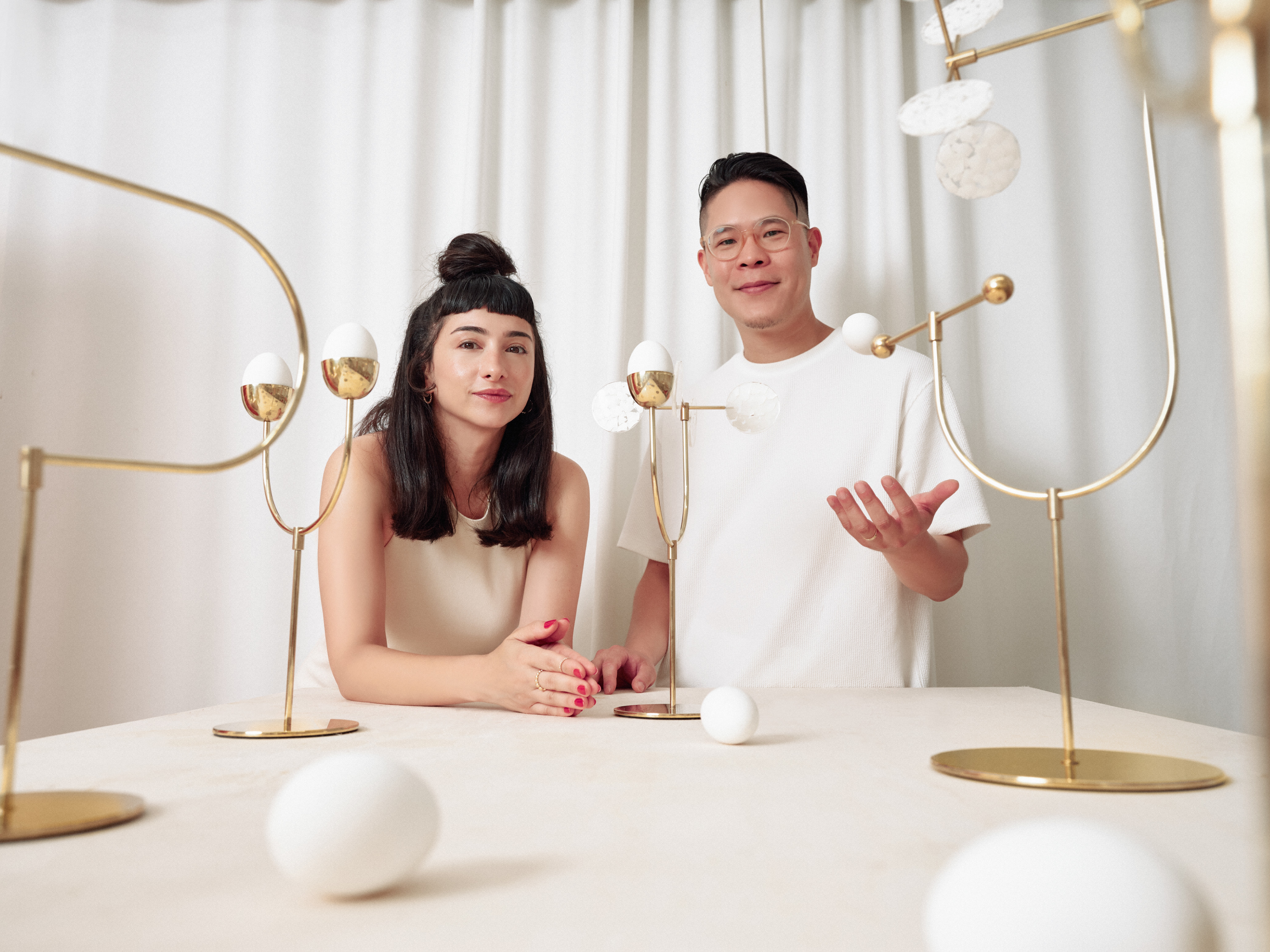 A brightly lit portrait of a young woman and a young man, both with dark straight hair, standing behind a table wearing neutrally tinted clothing before a series of golden candelabra holding eggs.