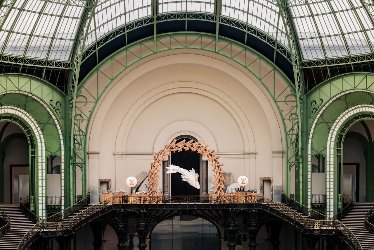 A gigantic fish-shaped sculptures floats in the air under a wooden arch in a green-hued, stunning glasshouse-like building.