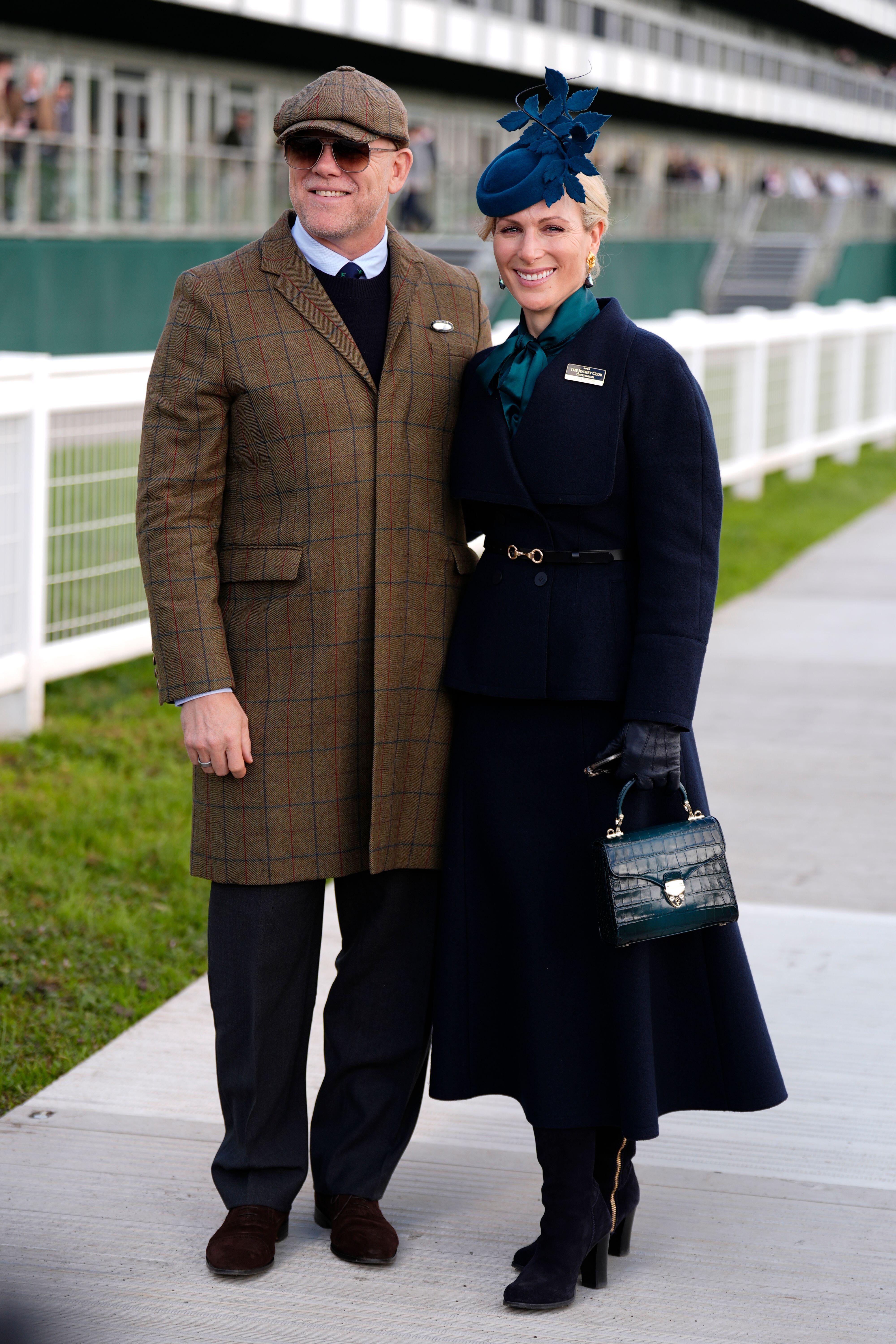 Mike Tindall wearing a flat cap and tweed coat standing next to Zara Tindall wearing a long blue coat and boots at Cheltenham races