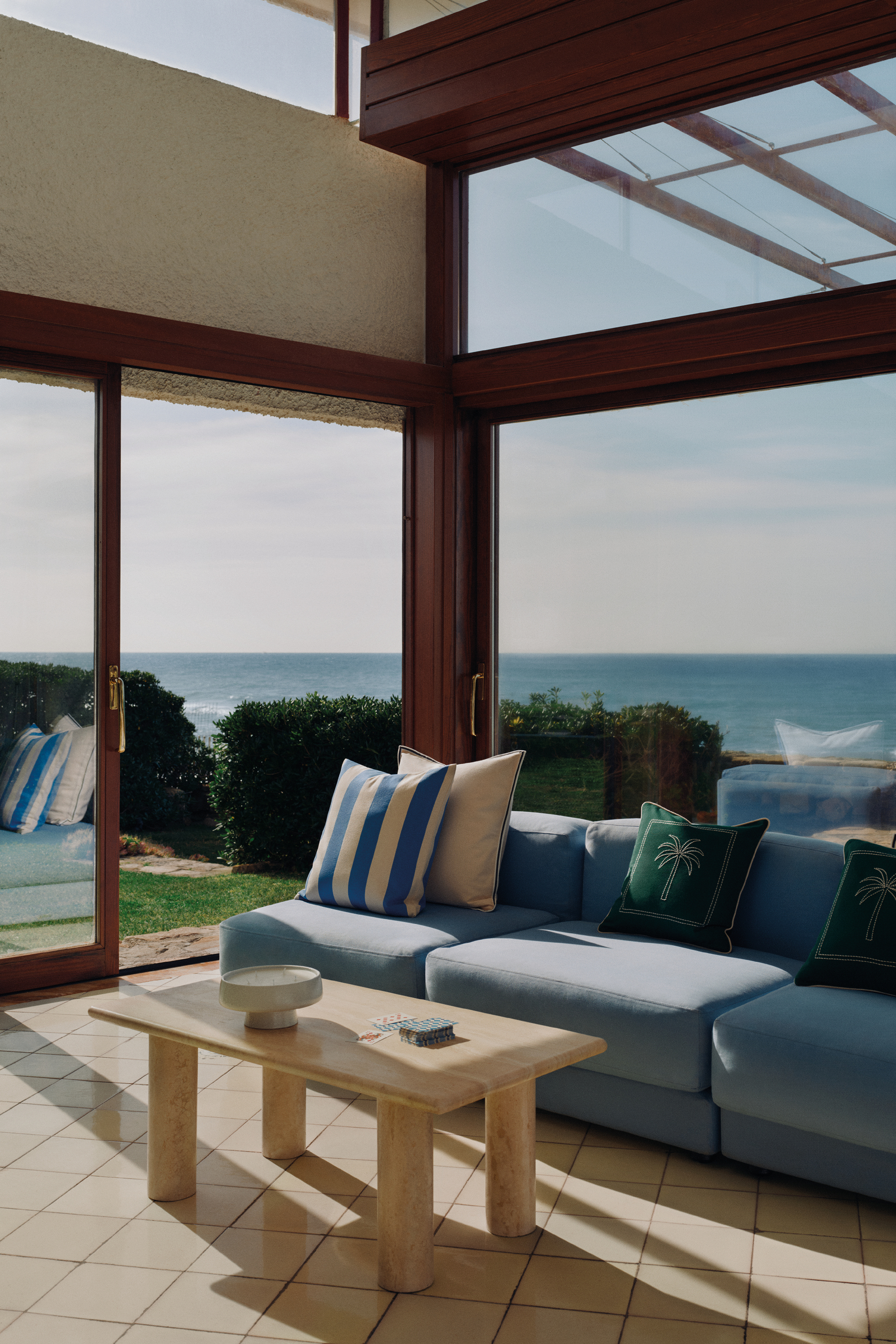 Image of a sunny room with lots of windows that show views of the sea. There is dark wood trim in the room, a light blue sofa, cream tiled floors, and a cream marble coffee table.