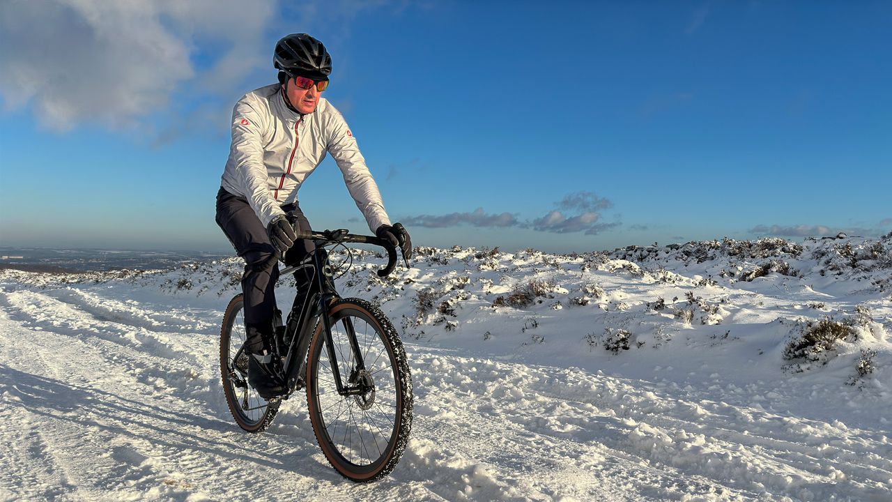 Man in a silver jacket riding a gravel bike on a snowy trail with blue sky