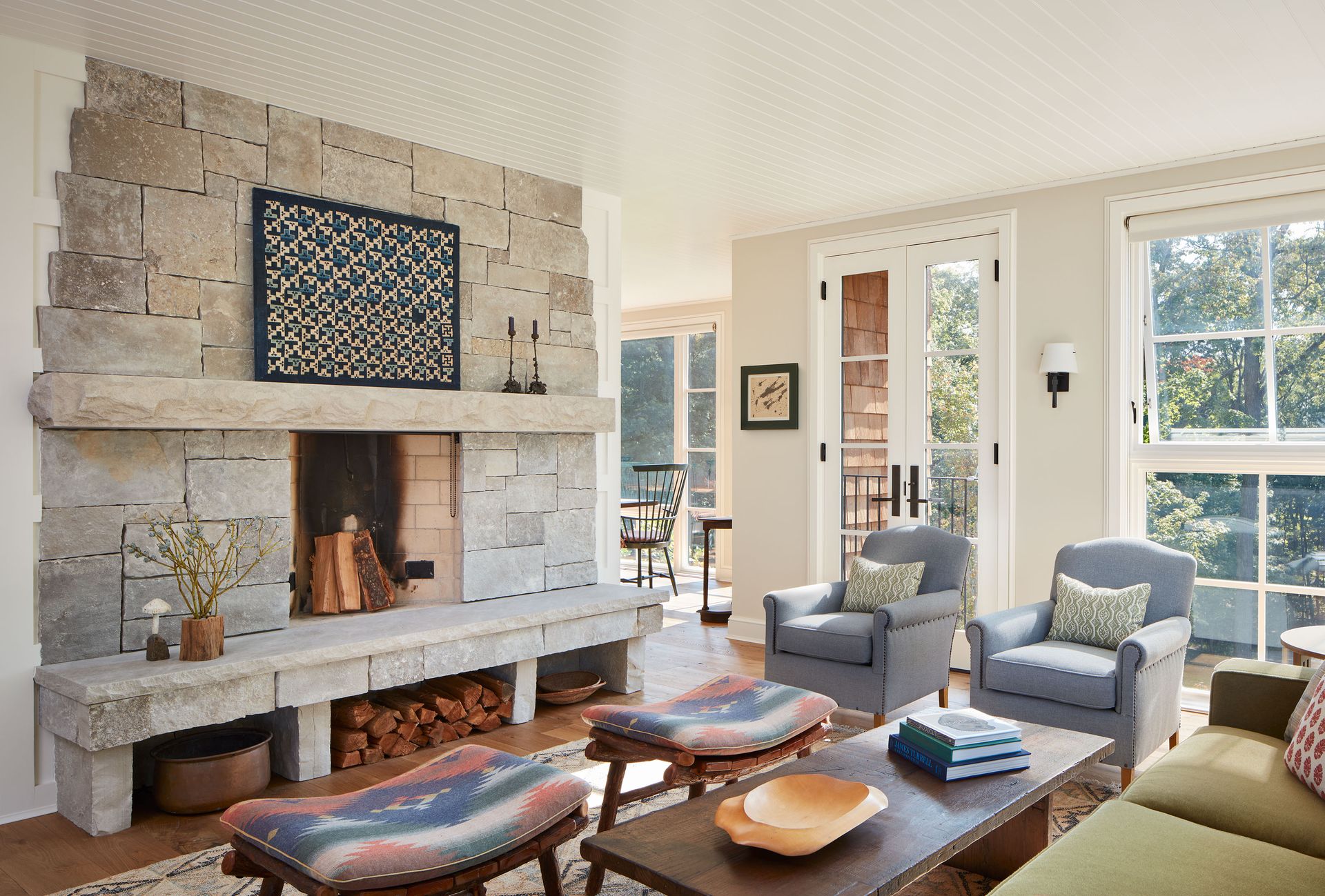 bright living room white paneled ceiling stone fireplace large windows green sofa pale blue armchairs twin kilim stools