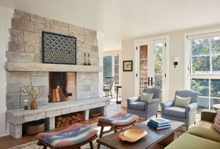 bright living room white paneled ceiling stone fireplace big windows green sofa pale blue armchairs twin kilim stools