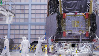 People wearing white coveralls and masks work around a giant covered piece of machinery in a cleanroom warehouse