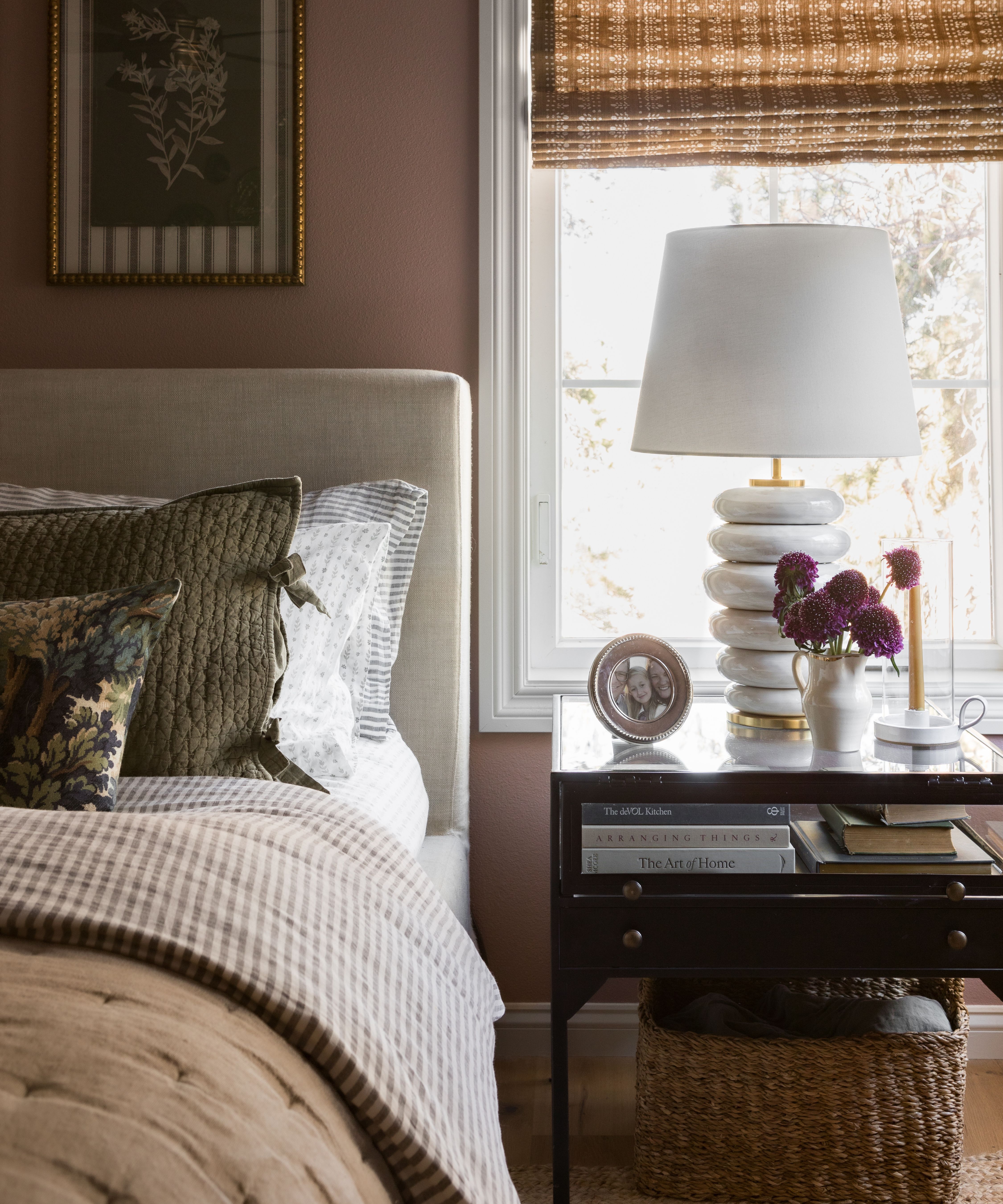 a dusky pink bedroom with a neutral linen bed, a window with a patterned blind, black nightstand with a white table lamp and layered striped bedding