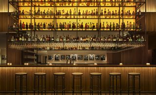 Alternative view of Bar Veraz at The Edition Barcelona hotel, Spain featuring a large high-level rack with multiple bottles of drinks and glasses with a dulled yellow light shining through. The bar design features vertical wooden slats and there are framed photos on a beige walls behind the bar and multiple bar stools in front
