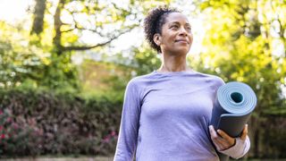 Woman smiling and walking through park holding yoga mat