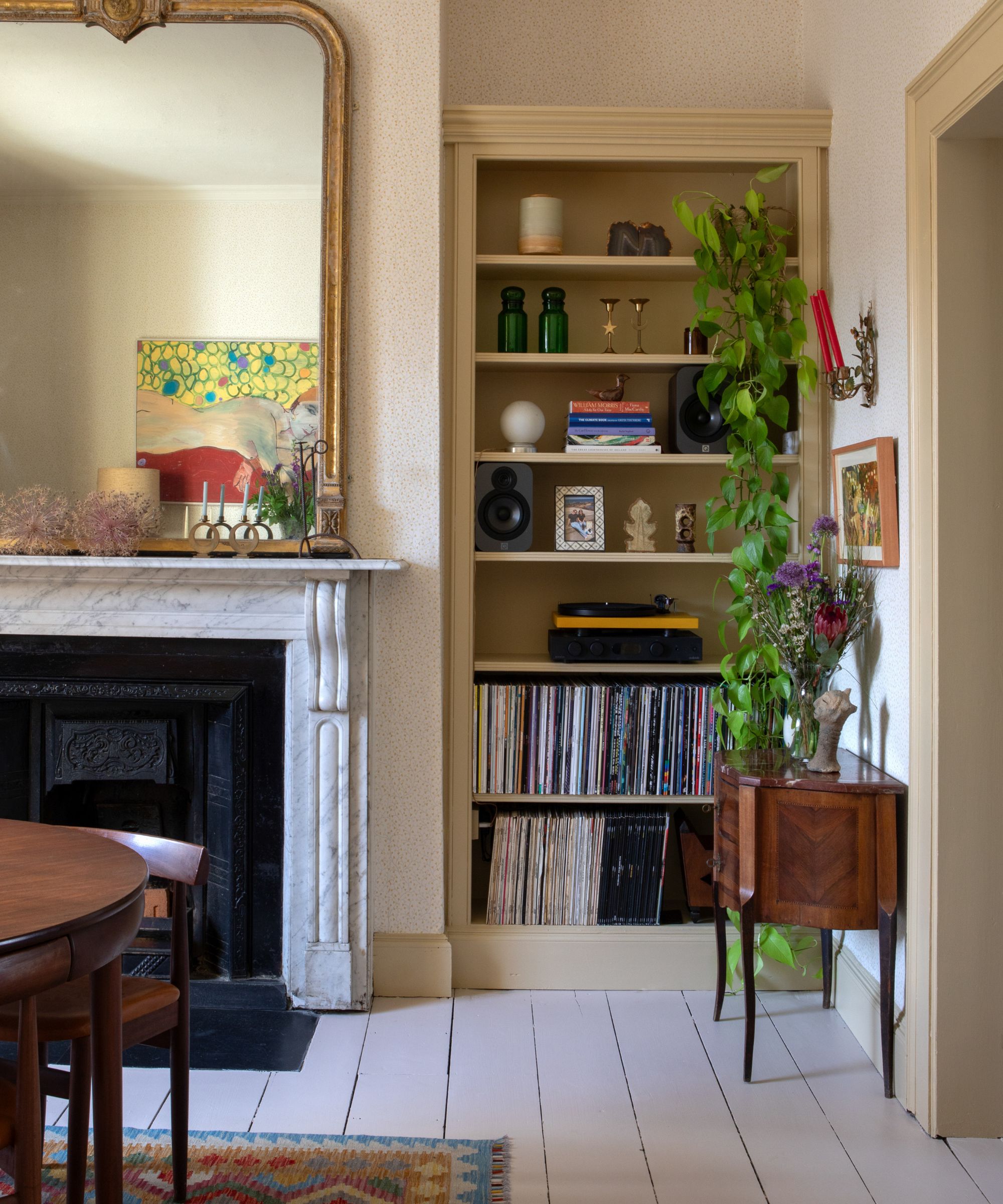 Dining room with painted wooden floor, a marble fireplace, a large gold mirror above, a built-in alcove with bookshelves decorated with books and trinkets, and ivy hanging down.