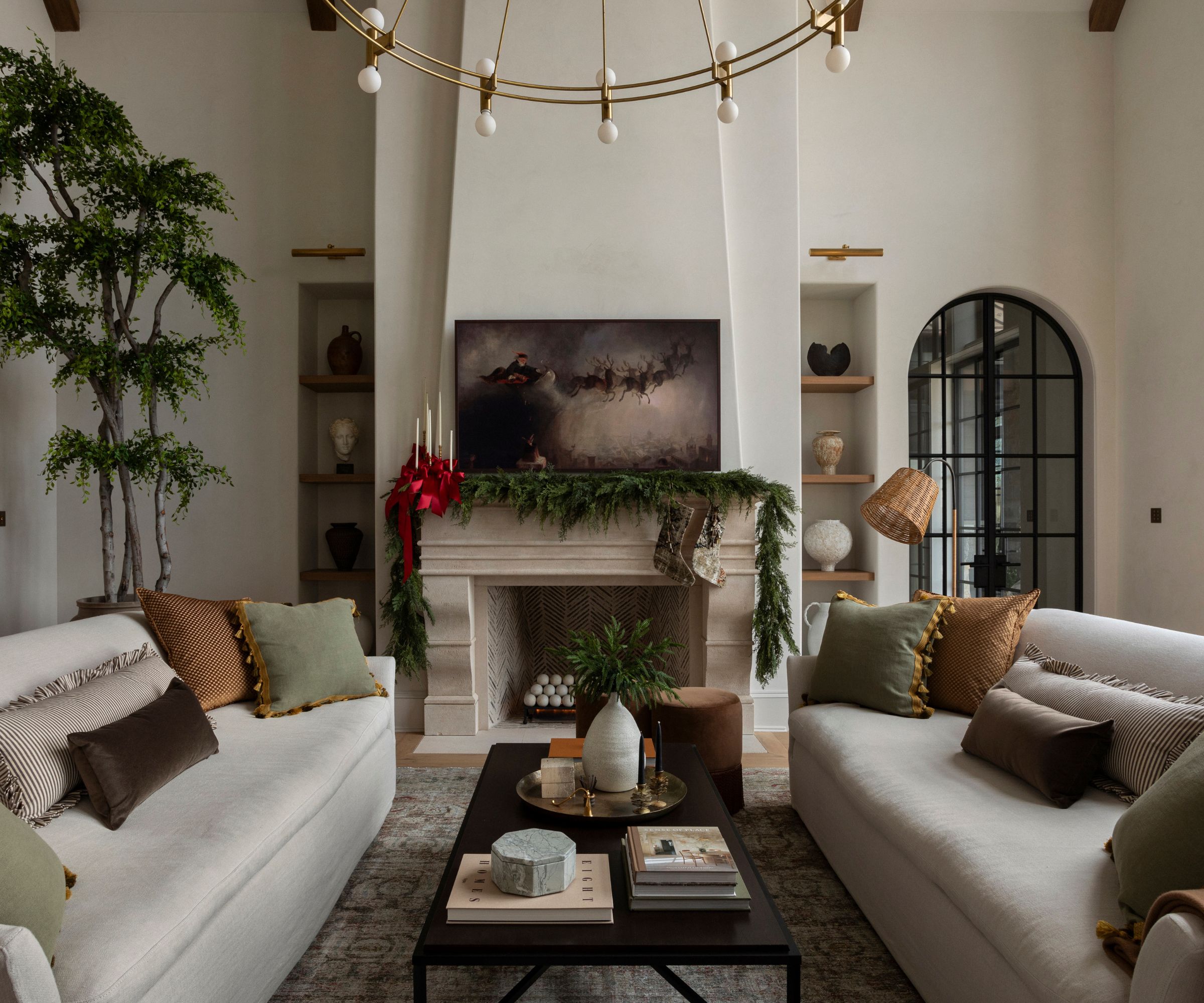A modern organic living room with a wreath hung along the mantel with stockings suspended and a group of candle holders wrapped with a red ribbon mixed into the holiday decor