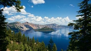 the annular solar eclipse will be visible from Crater Lake National Park, Oregon. Here the blue lake reflects the white clouds and is surrounded by tall evergreen trees.