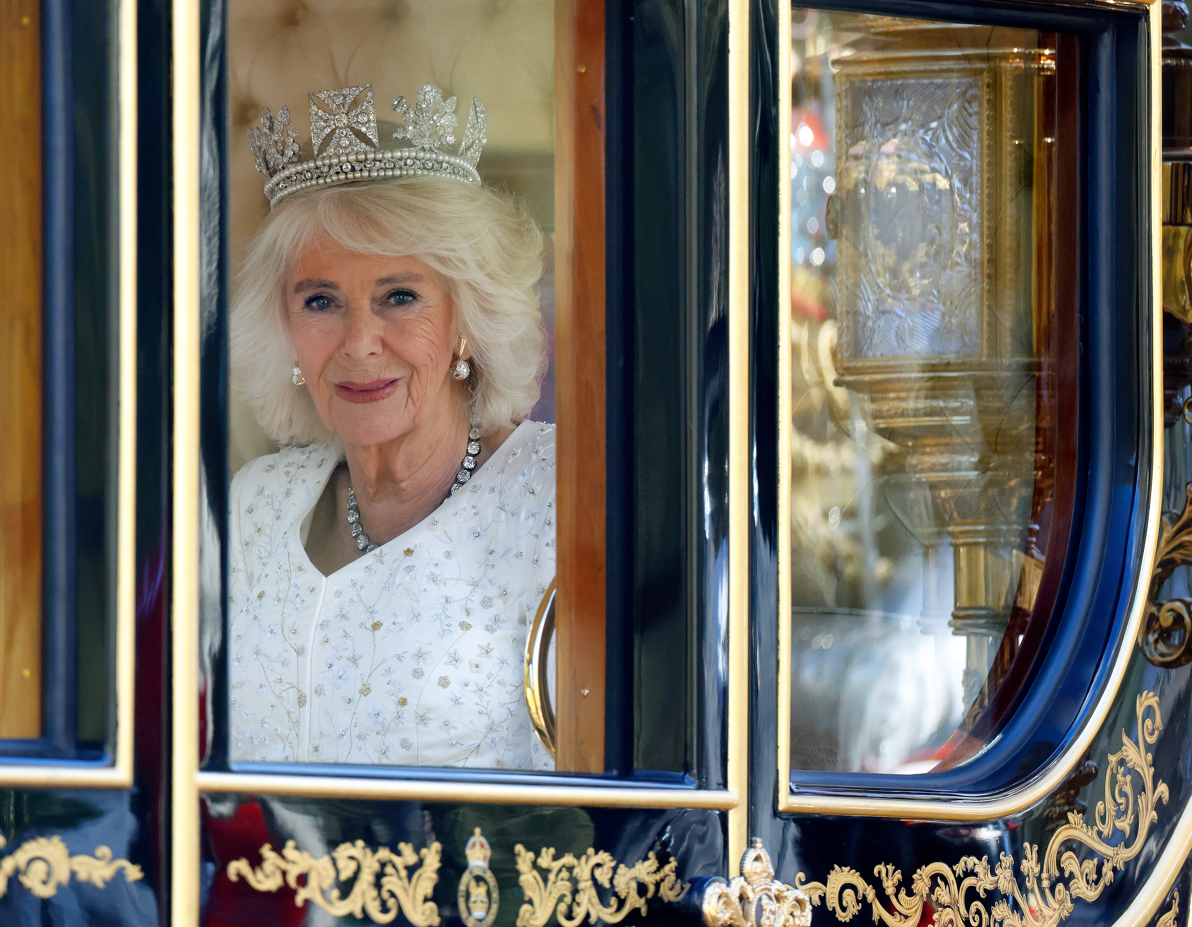 Queen Camilla wearing a crown and a white gown in a carriage