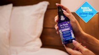 Woman holding a bottle of Dr Teal's Calm Blend Sleep Spray for Pillows and Bed Sheets next to two white pillows on a bed she is about to spray