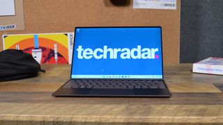 En Dell XPS 13 (2022) står öppnad på ett träbord med skrivbordet öppet på skärmen med en stor TechRadar-logo.