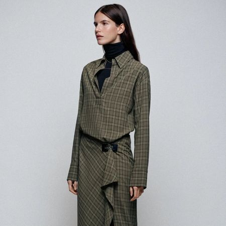 A close-up shot of a model wearing a black turtleneck top and green check midi dress with a belt from Zara