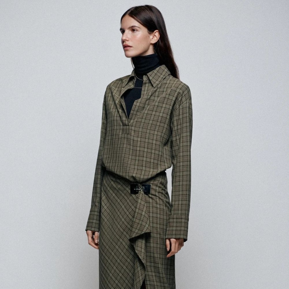 A close-up shot of a model wearing a black turtleneck top and green check midi dress with a belt from Zara