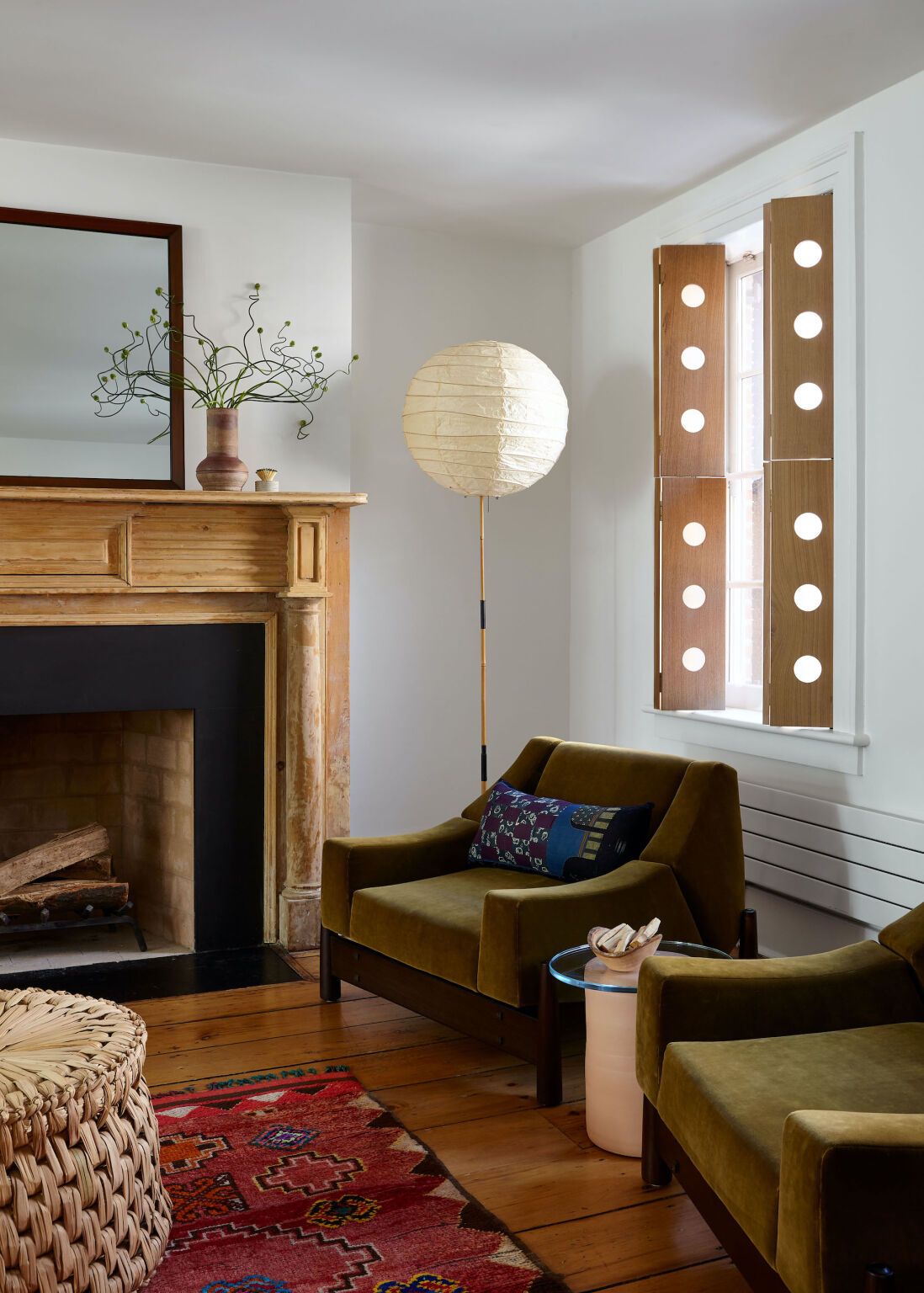 Cozy living room with olive green velvet armchairs, wooden fire surround, paper floor lamp and modern light-filtering shutters