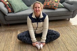 Kat Storr performs butterfly pose on a rug at home. She is sitting with her knees bent, feet touching and her back straight. She grasps her ankles as she performs the pose. Behind her we see a couch.