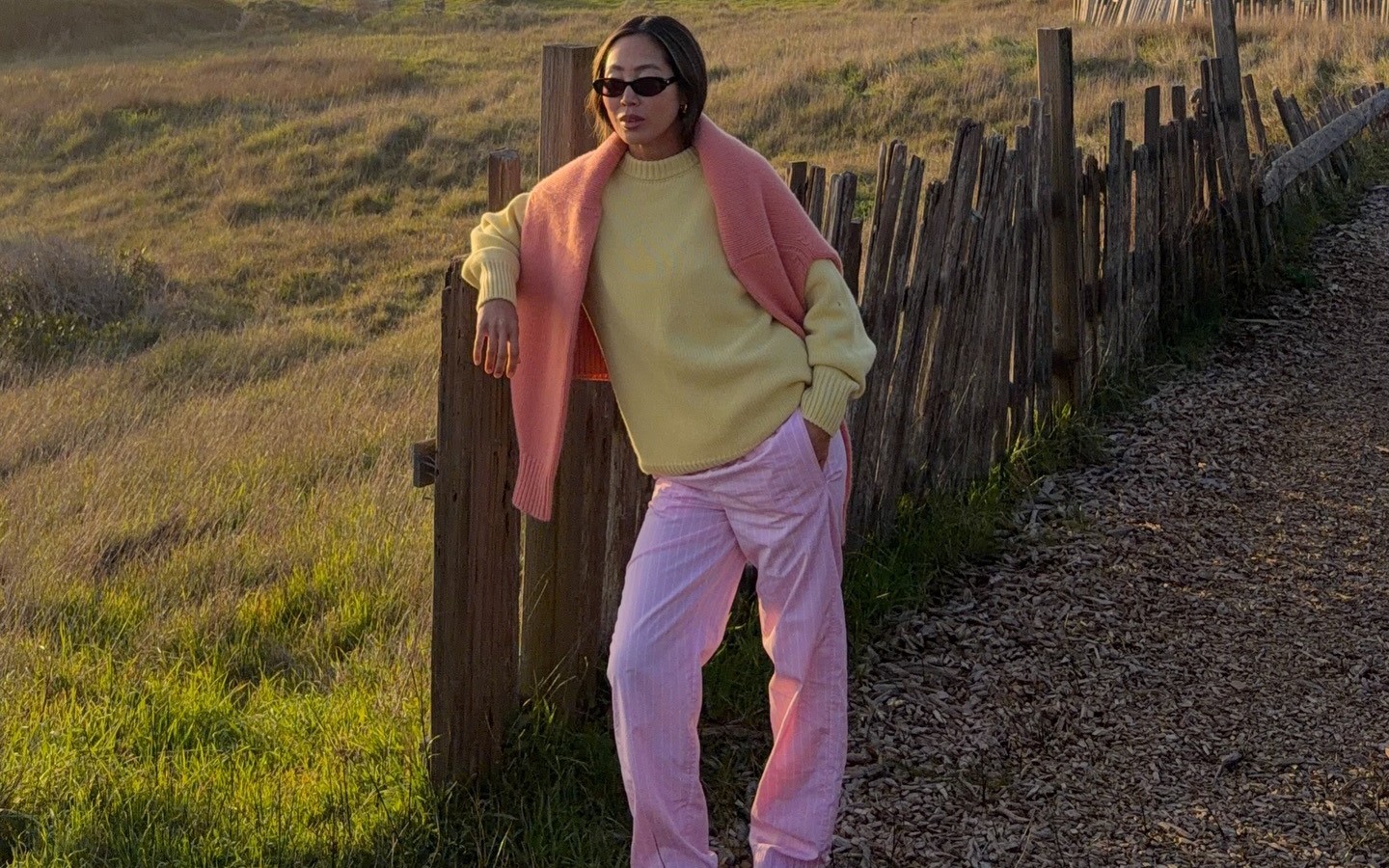 Aimee Song wears a pastel yellow sweater with a coral sweater draped over her shoulders and pink elastic waist pants in front of a sunset. 