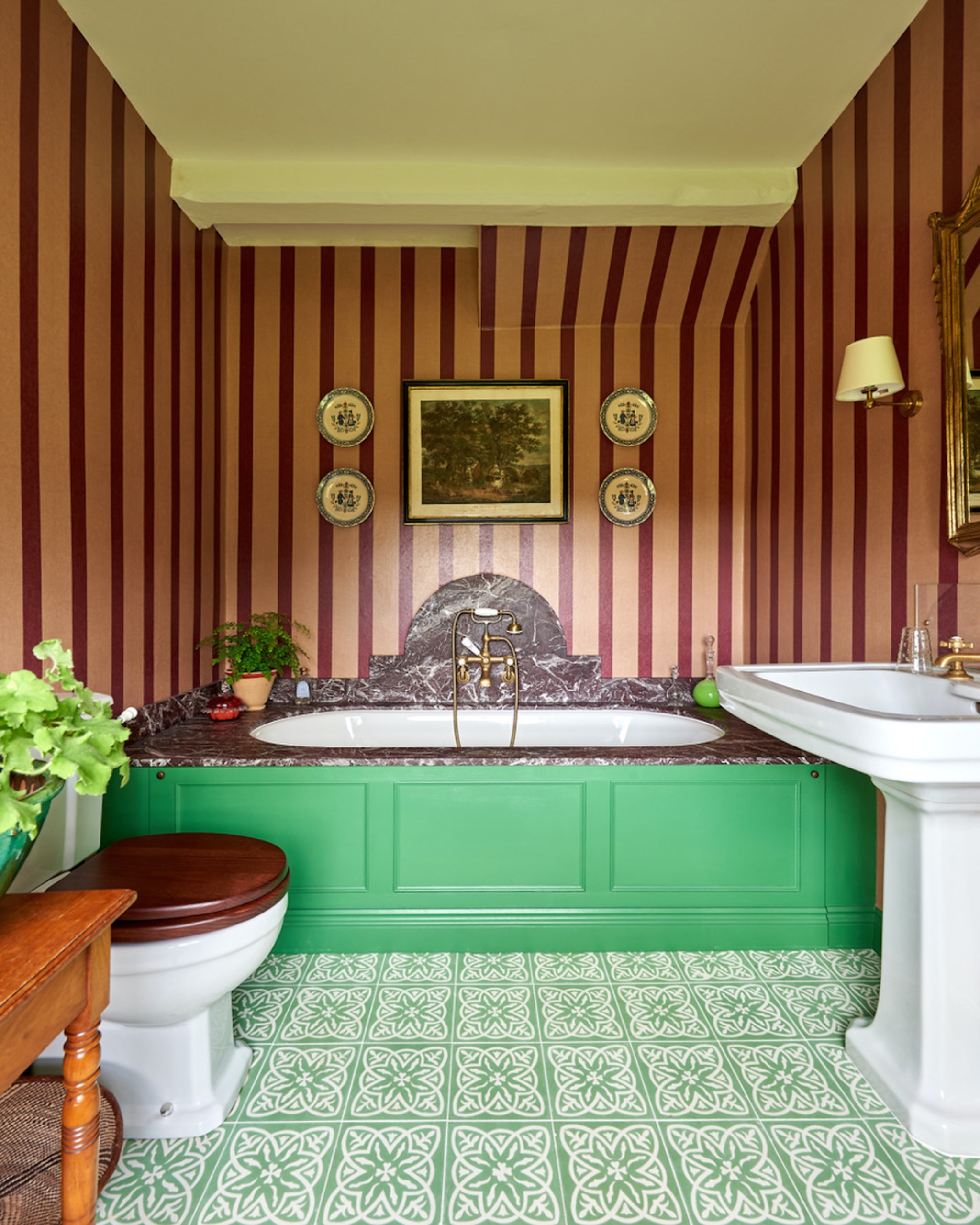 Bathroom with green-and-white patterned floor tiles, burgundy striped wallpaper, green bath paneling, a veined marble bath surround and a white pedestal sink.