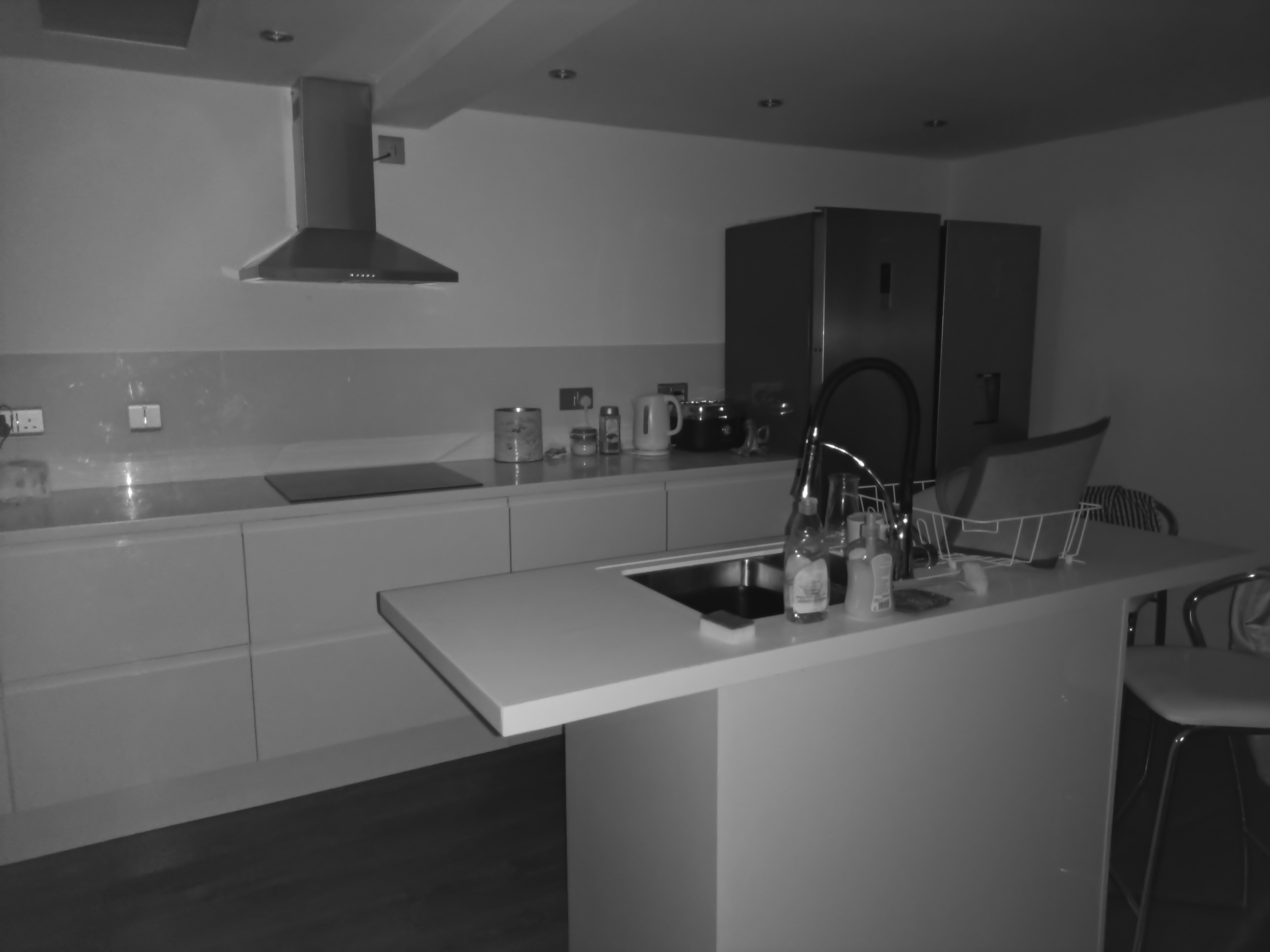 This black-and-white image shows a modern kitchen with a sink on a center island, stainless steel refrigerators, and a range hood.