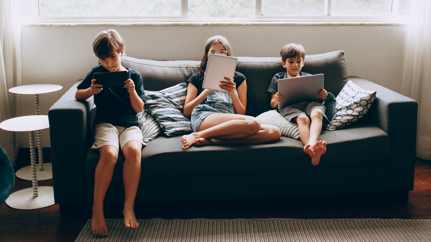 Three kids sitting on a couch playing with iPads and tablets