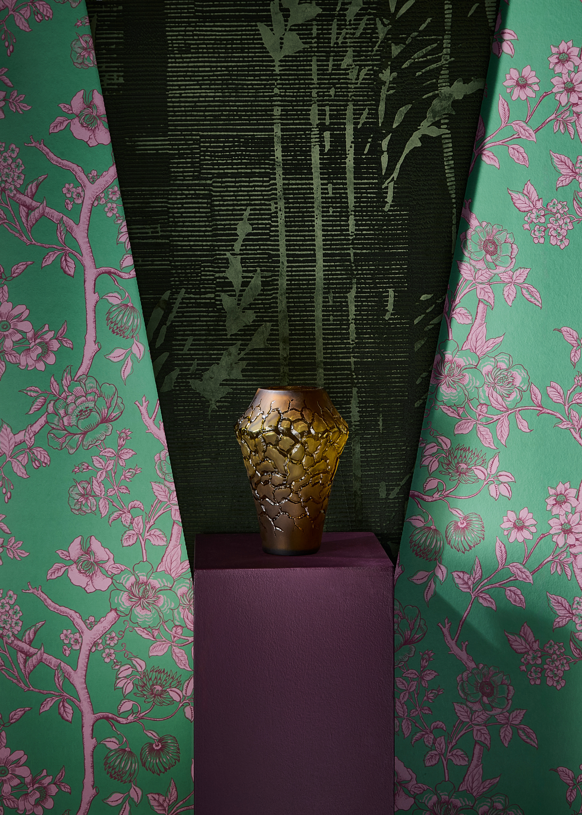 a design showcase featuring a purple pedestal with an ornate vase on top in a rust/gold almost cracked finish against a green and purple floral patterned wallpaped with a darker green material over the top in the centre pointing down towards the back of the vase