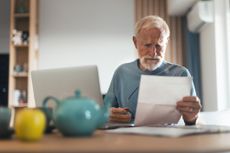 Pensioner looks at documents as he sits at desk beside laptop.