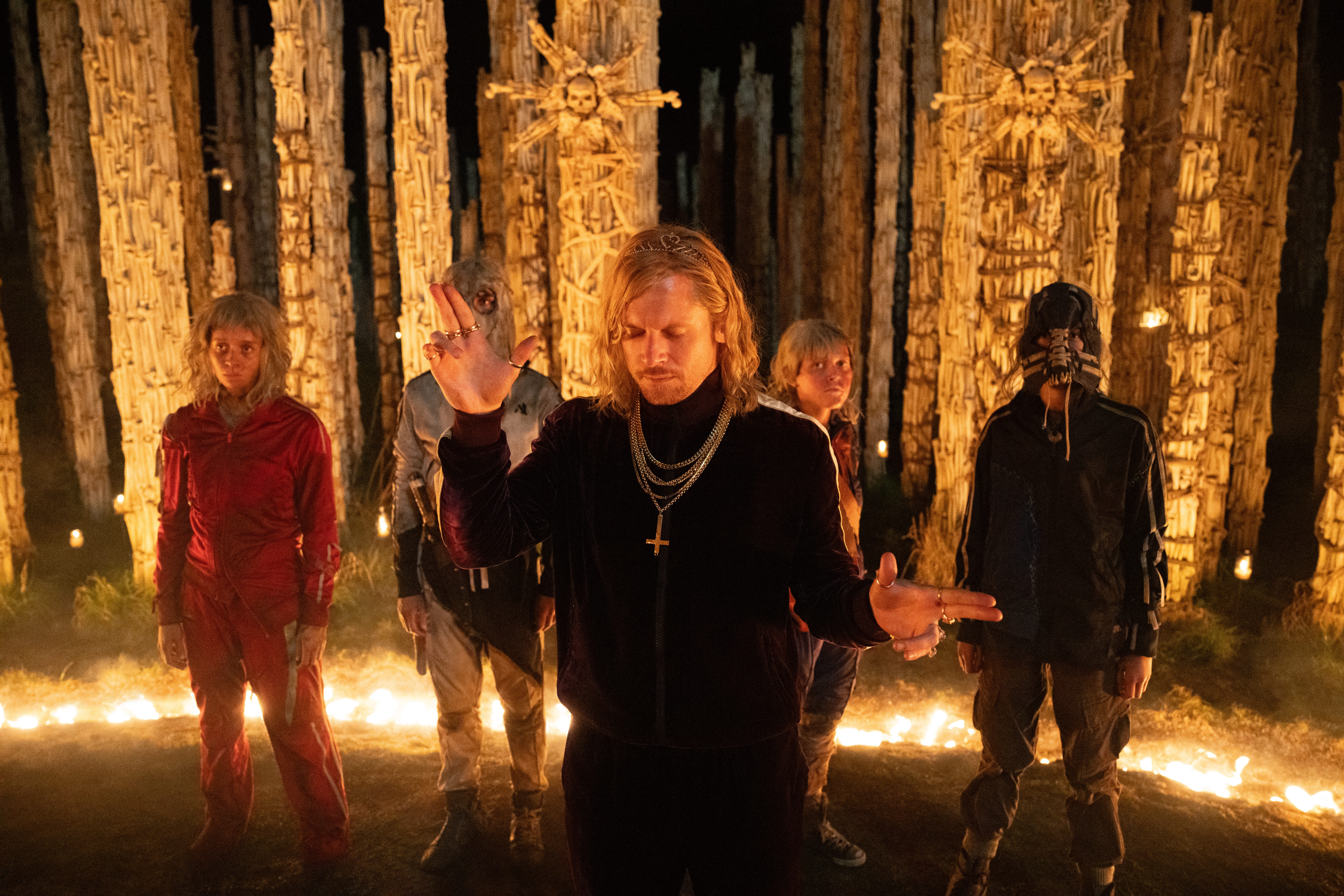 the jimmies including one played by jack o'connell in blonde wigs standing around a ring of fire and the yellow lit bone temple in a still from 28 years later the bone temple