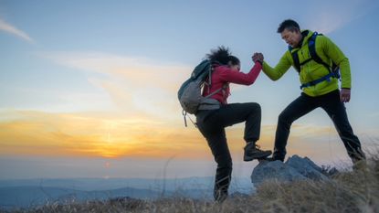 Side view of man helping woman while ascending mountain against sky at sunset.